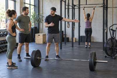Coach briefs members beside a loaded barbell, pull-up rig and fan bike in a functional training gym